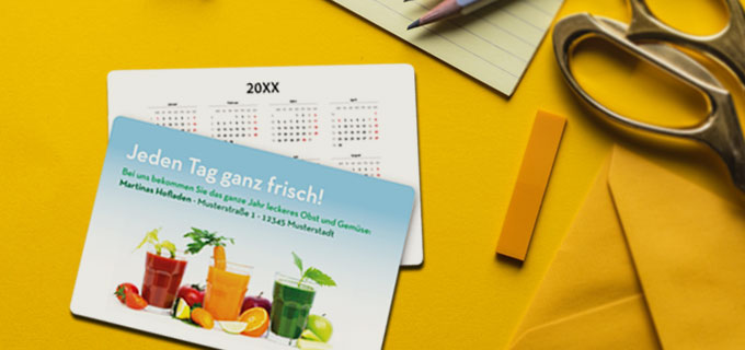 Zwei bedruckte Taschenkalender liegen auf einem gelben Schreibtisch. Die Minikalender mit Logo zeigen drei Gläser mit Gemüsesäften. Auf den bedruckten Kalendern steht "Jeden Tag ganz frisch!".
